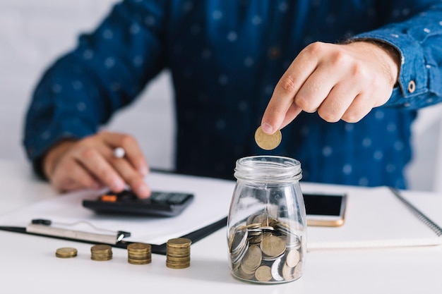 Close-up of man's hand putting coin in jar using calculator