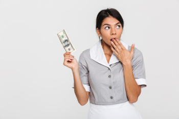 Portrait of amazed attractive maid in uniform holding hundred dollars while covering her mouth with hand, looking aside Free Photo