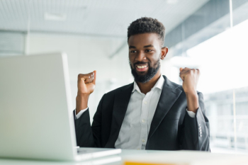 Happy african-american businessman in suit looking at laptop excited by good news online. black man winner sitting at office desk achieved goal raising hands celebrating business success win result Free Photo