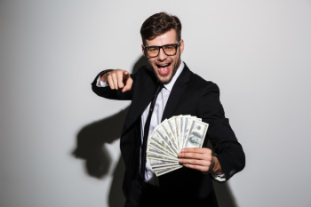 Happy attractive man in glasses and black suit holding bunch of money while pointing with finger on you Free Photo