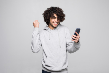 Young curly man playing with the phone looks happy and fisted after winning, isolated on a white wall. Free Photo
