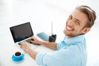 Young handsome cheerful confident businessman sitting at table working on laptop with cup of coffee aside.  smiling. white modern office interior Free Photo