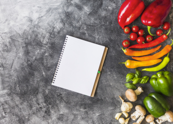 An overhead view of blank spiral notebook with colorful vegetables on rough background