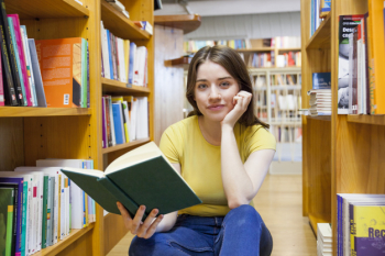 Teen girl with book looking at camera between bookshelves