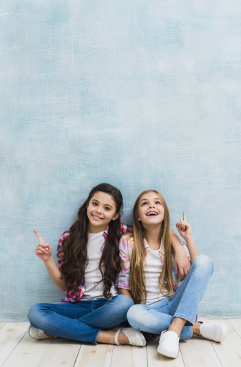 Happy two girls sitting in front of blue wall pointing their finger upward