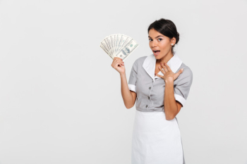 Portrait of amazed young housekeeper in uniform holding fan of dollar banknotes Free Photo