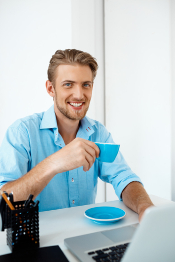Young handsome cheerful confident businessman sitting at table working on laptop drinking coffee.  smiling. white modern office interior Free Photo