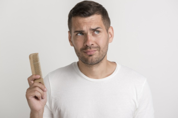 Confused young man holding comb looking away standing against white wall Free Photo