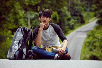 a handsome hiker is sitting on a road and putting hand on his chin