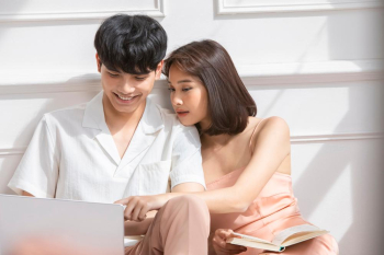A handsome young man is using a laptop while his wife is reading a book