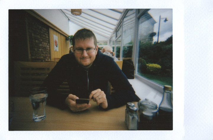 analog,cellphone,eatery,eyeglasses,film photography,holding,indoors,instant film,looking,male,man,mobile phone,polaroid,restaurant,sitting,smartphone,smile,smiling,table,waiting